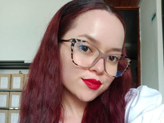 NaomyRose met rode lippenstift, een bril en rood haar, die lichtjes een frons trekt, draagt een witte blouse en laat haar borsten subtiel zien. Haar achterkant is niet zichtbaar. Ze kijkt naar de camera..