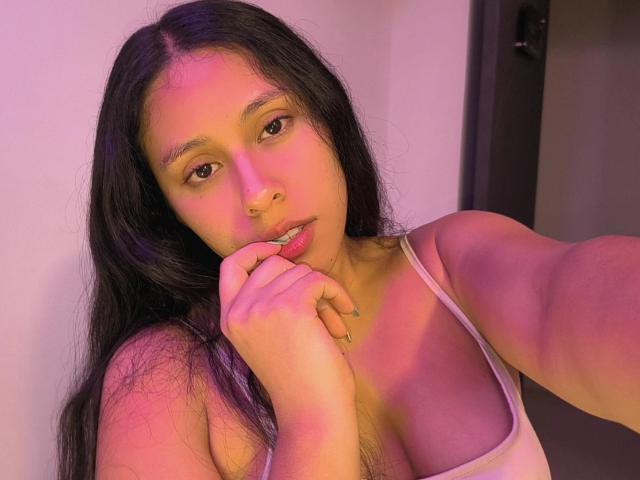 MarilynHills with long black hair, pinkish-purple lighting, white tank top, biting her finger, showing her tits, and a nose ring, looking at the camera.