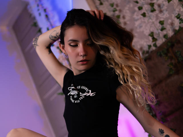 LilaLaurent with long blonde and dark brown wavy hair, wearing a black tee, has tattoos on her arm, and is sitting with one hand in her hair, looking down.