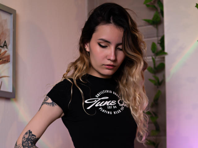 LilaLaurent has wavy blonde and dark brown hair, wearing a black T-shirt, looking down with subtle makeup, tattoos on her arm, and a cute, sexy, and attractive face.