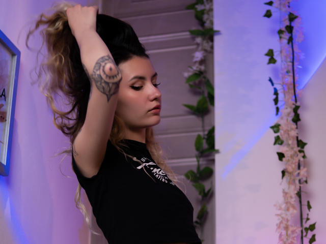 LilaLaurent with long, wavy blonde hair, black t-shirt, tattoo on right forearm, holding hair up, side profile, blue and purple lighting, green leafy decoration, subtle makeup.