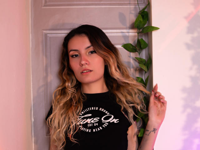 LilaLaurent with long, wavy blonde and brown hair, wearing a black shirt, standing against a door, holding her hair with a green vine on the right.