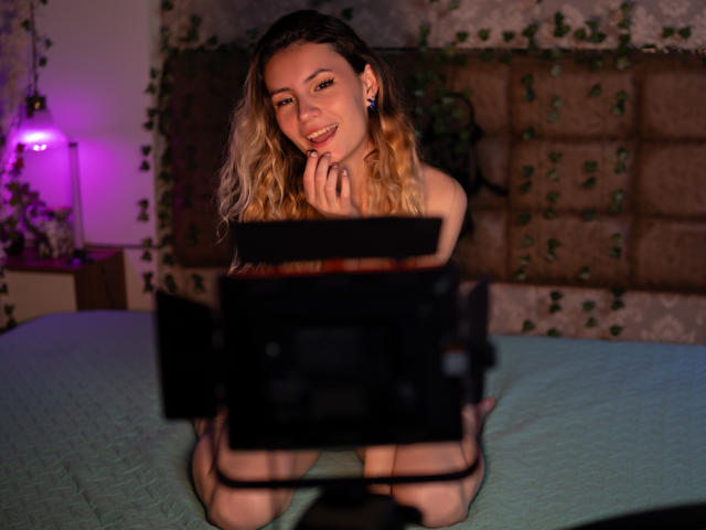 LilaLaurent, blonde, wavy hair, smiling, kneeling on bed, green quilt, naked, holding camera, camera in foreground, purple light, vine-patterned wall, cute, sexy, small tits, pink nipples.
