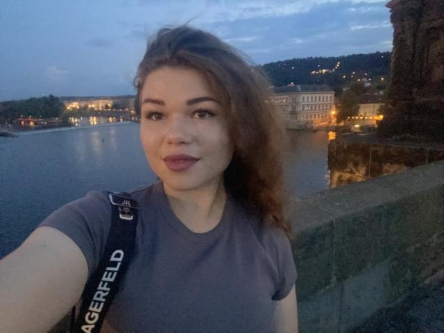 Miali69 mit braunen, welligen Haaren, einem grauen T-Shirt, einem schwarzen Rucksack, lächelnd in die Kamera, Dämmerung am Wasser, dezentes Make-up, natürliches Licht, ruhiger Gesichtsausdruck, sanftes, blaues Abendlicht, ungezwungenes Selfie..