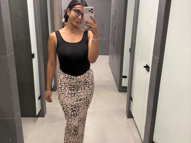 SarahPrincess with glasses, black tank top, and leopard-print skirt, holding phone, smiling, showing her ass in a gray-tiled hallway.