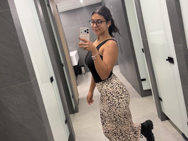 SarahPrincess with glasses, black tank top, and leopard-print pants, holding a phone in a bathroom, showing her curvy ass and small tits, smiling at the mirror.