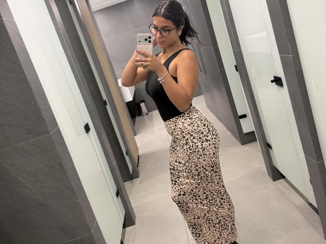 SarahPrincess with black tank top, leopard print skirt, glasses, and medium tits, taking a mirror selfie in a gray-tiled room, holding a pink phone with a gold case.