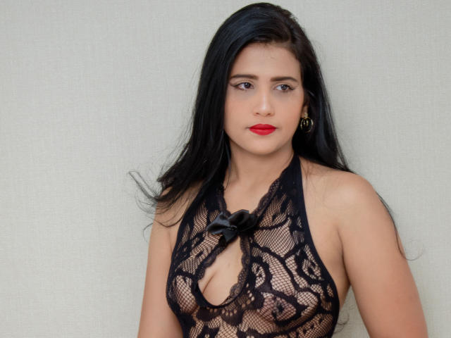 XAshleyRoseX trägt eine schwarze Spitzenbluse, die ihre Brüste zeigt, hat roten Lippenstift, langes schwarzes Haar und goldene Creolen und steht vor einer schlichten Wand..
