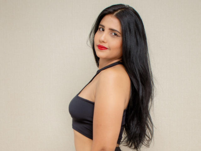 XAshleyRoseX avec de longs cheveux noirs, du rouge à lèvres rouge et un soutien-gorge de sport noir, montrant ses seins, regardant par-dessus son épaule, souriant..