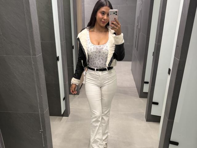 SarahPrincess mit durchschnittlicher Brustgröße, trägt eine schwarze Jacke mit Fellbesatz, ein weißes Oberteil mit Blumenmuster und weiße Hosen, macht ein Selfie in einem grauen Badezimmer mit Fliesen und hält ein pinkes iPhone..
