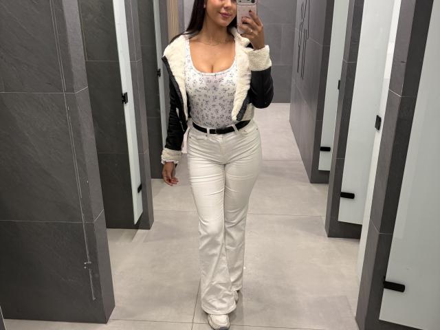 SarahPrincess, con un seno prosperoso, indossa una giacca nera e bianca con fodera in pelliccia, pantaloni bianchi e una canotta bianca aderente, e sta scattando un selfie allo specchio in un negozio..