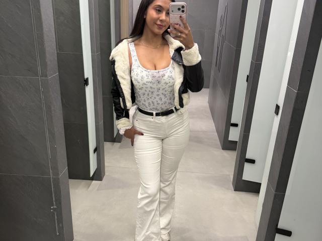 SarahPrincess mit durchschnittlicher Brustgröße, trägt ein blumiges Oberteil, eine schwarze Jacke, weiße Hosen und weiße Schuhe und macht ein Selfie vor einem Spiegel in einer Umkleidekabine, wobei sie ein rosa Handy hält..