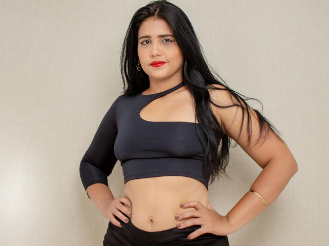 XAshleyRoseX has long black hair, red lipstick, and a dark blue, cut-out, long-sleeve crop top, showing her tits and a flat stomach, with hands on her hips.