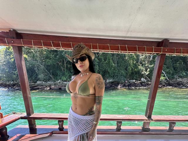 FerrazJuanitta69 met grote borsten, in een groene bikini-top, een witte kanten rok, een cowboyhoed en zonnebril, staand op een boot met groen water en een bos op de achtergrond..