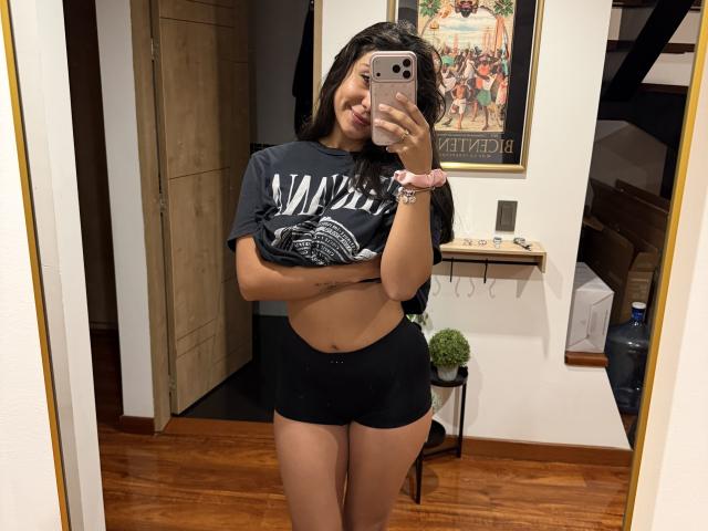 SarahPrincess with tan skin, black shorts, and a lifted black shirt showing her small tits, taking a mirror selfie, smiling, wearing a pink scrunchie, holding a rose gold phone.