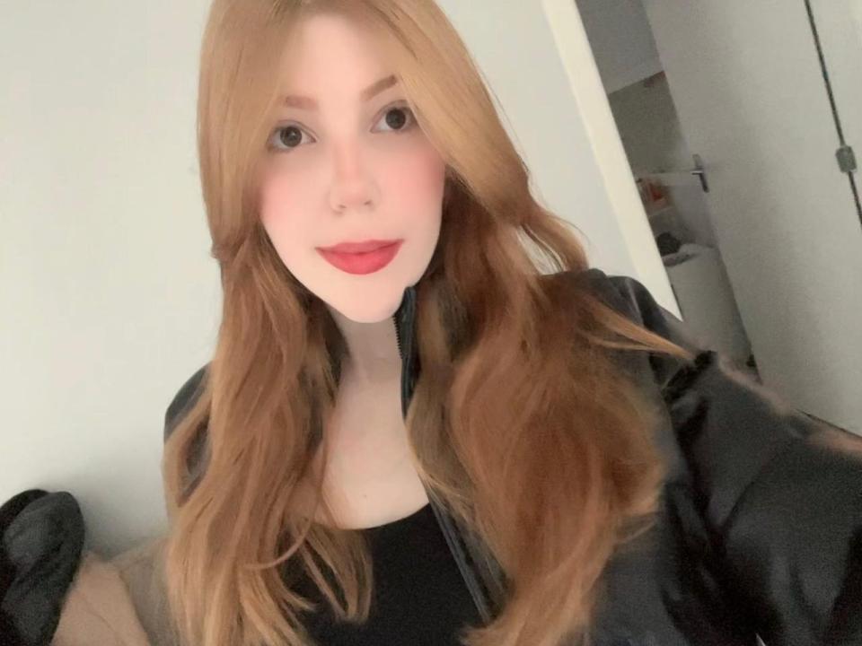 CloeZinhaX hat langes, welliges, rotes Haar, roten Lippenstift und trägt eine schwarze Lederjacke. Sie hat helle Haut und ein leichtes Lächeln, auf einem Selfie..