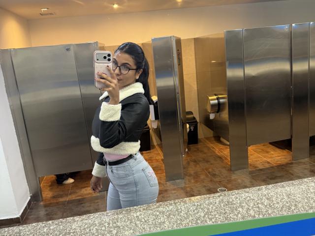 SarahPrincess with glasses, black fur-trimmed jacket, pink top, and tight light-blue jeans, taking a mirror selfie in a bathroom, showing her curvy ass and small tits.