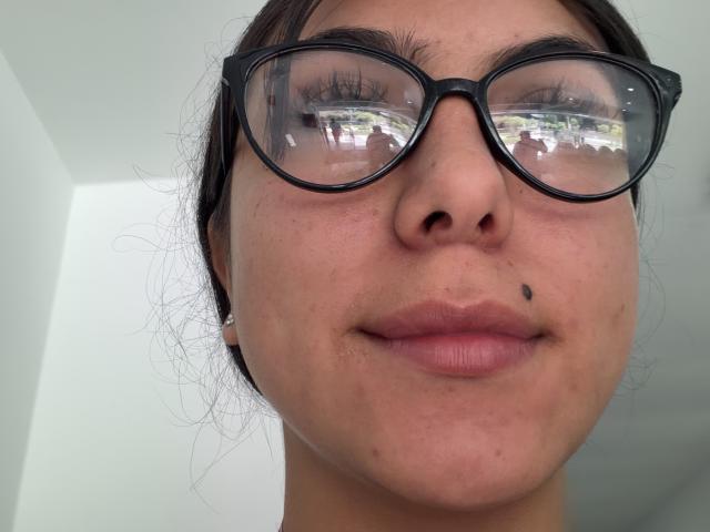 SarahPrincess has a mocha skin tone, black-rimmed glasses, and a small black mole on her right upper lip. She's wearing a pearl earring and has a slight smile.