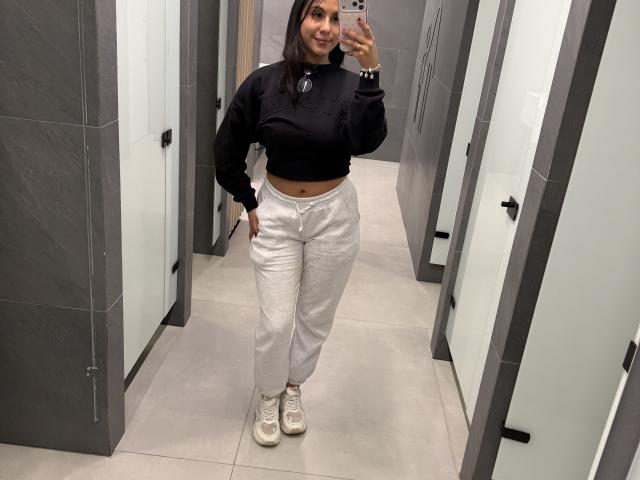 SarahPrincess with long dark hair, black hoodie, white pants, and white sneakers, smiling in a mirror selfie, showing her midriff and hand in pocket.