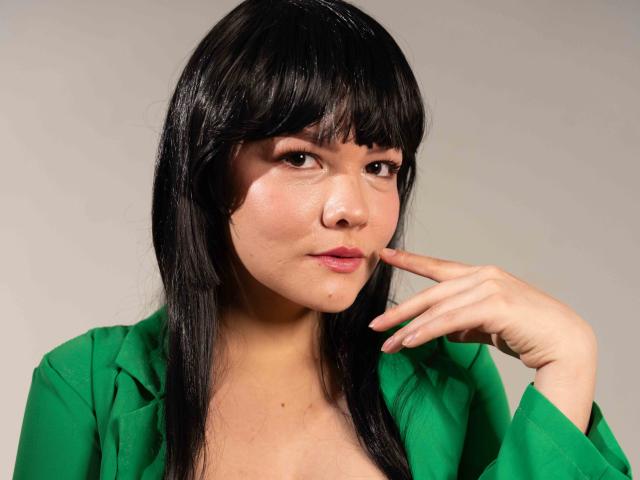 Amaranta Soto tiene el pelo negro, con flequillo, y lleva una camisa verde. Está tocándose la barbilla con un dedo, y parece un poco confundida. Sus pechos son pequeños, y tiene una cara bonita..