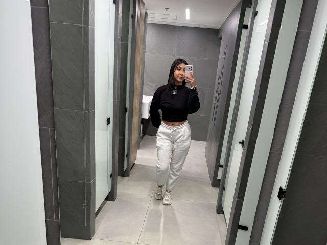 SarahPrincess standing in a bathroom, wearing a black crop top, white satin pants, and beige sneakers, taking a mirror selfie with her phone.