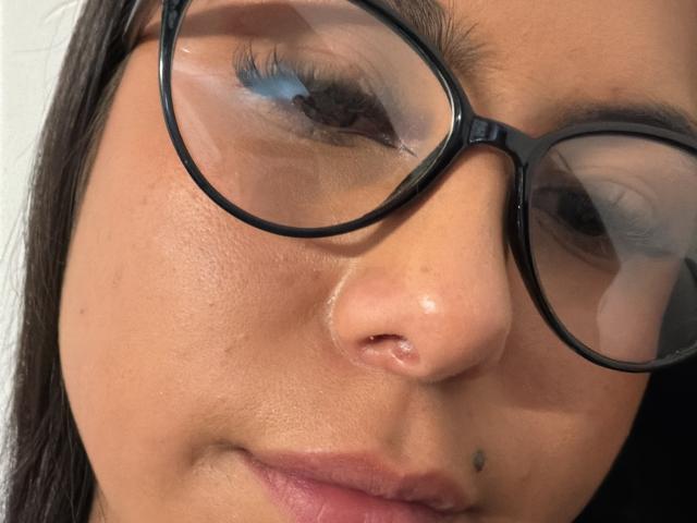SarahPrincess's face, wearing black glasses, with blue eyeshadow, a small black mole near her lower lip, and a slight nose piercing, showing a calm, slightly smiling expression.