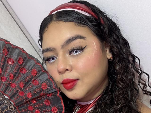 AprielKeith has curly black hair, red lipstick, and a red headband, holding a red and black lace fan, with winged eyeliner and a small scar on her cheek.