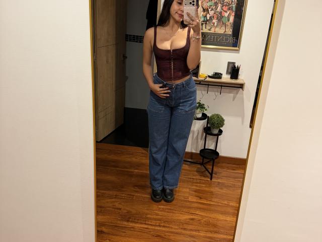 SarahPrincess with big tits in a burgundy corset, blue jeans, and black shoes, taking a mirror selfie, showing off her curvy, cute, and sexy figure.