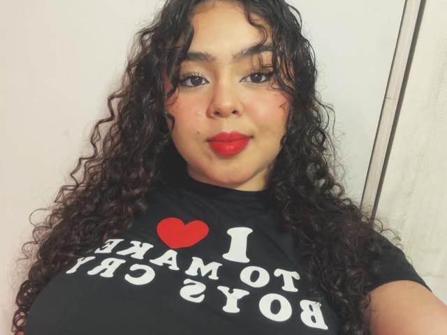 Apriel Keith, met krullend donker haar, rode lippenstift en een zwart shirt met een hart en tekst, heeft grote borsten en een lichte glimlach, en staat tegen een witte muur..