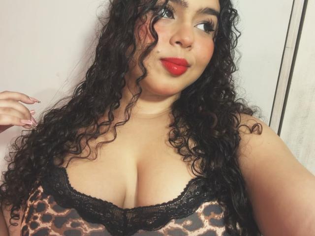 Apriel Keith, met krullend zwart haar, rode lippenstift en een top met luipaardprint die haar grote borsten laat zien, kijkt opzij met een lichte glimlach, heeft roze nagels en houdt haar haar vast..