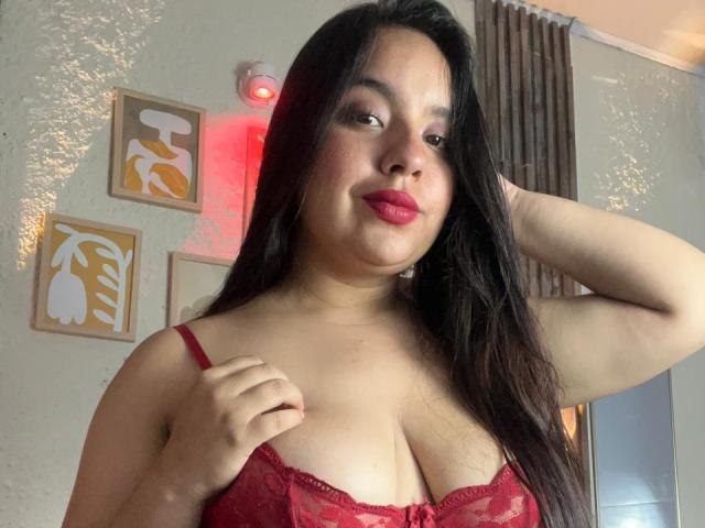 SammyTitss, a curvy Latina woman with long dark hair, red lipstick, and red lace bra, holding her bra strap, showing her large, rounded tits.