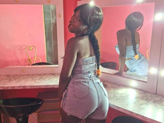 MissDominik with dark skin, long braided hair, wearing a strapless denim romper, showing her ass in a mirror, surrounded by lit bulbs. Reflection highlights her back.