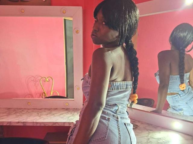 MissDominik with dark skin, long braided hair, wearing a strapless denim dress, showing her back and butt, standing in front of a mirror with a pink wall.