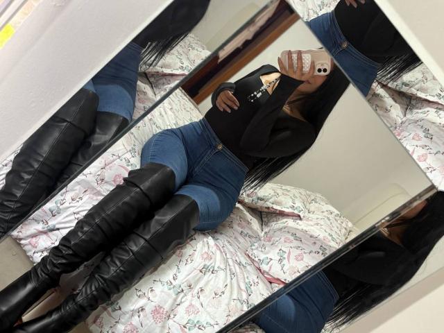 AntonellaPreschot with long black hair, wearing a black sweater, blue jeans, and black knee-high boots, taking a mirror selfie, showing her ass in the reflection.