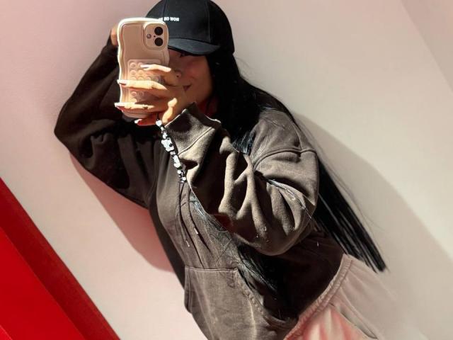 AntonellaPreschot with long black hair, wearing a black cap, brown leather jacket, and pink pants, taking a mirror selfie with a gold phone, showing a cute star tattoo.