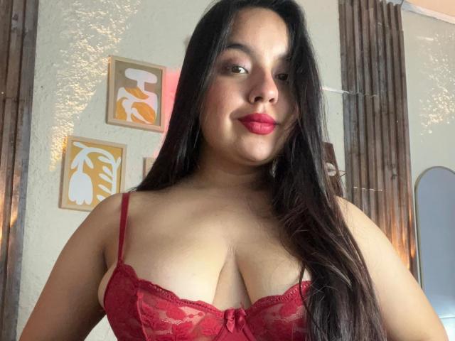 SammyTitss with long dark hair, red lace bra showing her big tits, pink lipstick, and a slight smile, standing in a room with wooden doors.