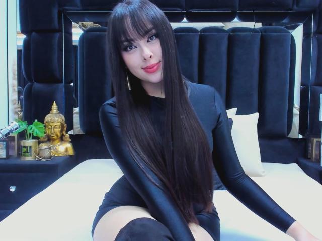 Alison Tatcher, avec ses longs cheveux noirs, son rouge à lèvres rouge, son haut noir à manches longues et ses bas noirs montants jusqu'aux cuisses, assise sur un lit, montrant son joli visage et ses fesses lisses..