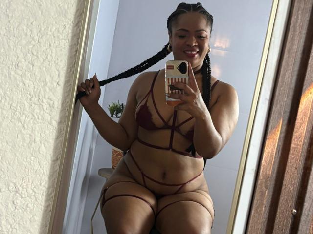 AliceBooty in maroon strappy lingerie, holding braided ponytail, smiling, showing her curvy, dark-skinned body and large, round ass in mirror selfie.