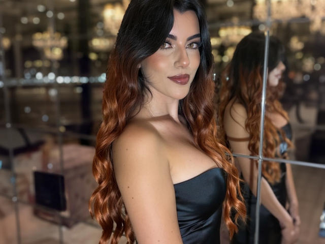 ZoeeMar with long, wavy, brown-to-red ombre hair, wearing a strapless black dress, standing in front of a mirror, showing off her ass and a sexy smile.