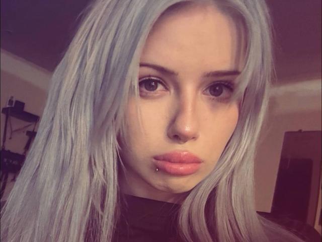 Anabel Tanberg, com longos cabelos prateados, lábios rosados e um lábio inferior com piercing, olhando diretamente para a câmera com uma expressão séria, sem que seus seios sejam visíveis..