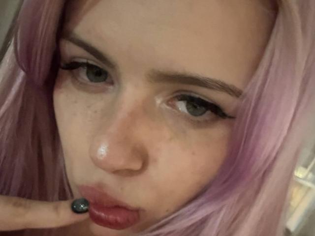 Anabel Tanberg hat rosarote Haare, blaue Augen und zieht eine Grimasse, wobei ihr Finger ihre roten Lippen berührt. Sie hat Sommersprossen und schwarzen Eyeliner. Ihre Nase ist leicht glänzend..