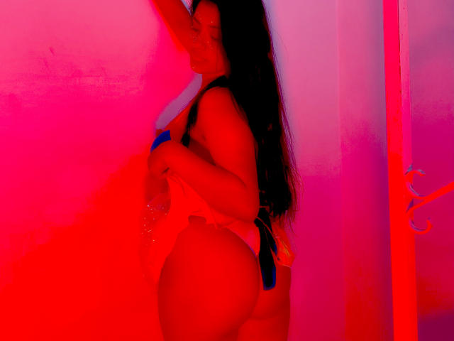 ArianithaXox with long black hair, standing sideways, holding a blue dildo against a red-pink wall, wearing a strap-on, showing her big ass, in a neon-lit room.