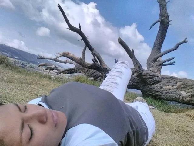 ArianithaXox with eyes closed, lying on grass, wearing grey and blue shirt, white pants, and white shoes, with a large, leafless tree in the background.