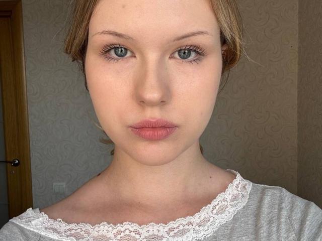 EllamaeRaugust has pale skin, blue eyes, and pink lips with a serious expression. She's wearing a white lace-trimmed top, and her brown hair is pulled back.
