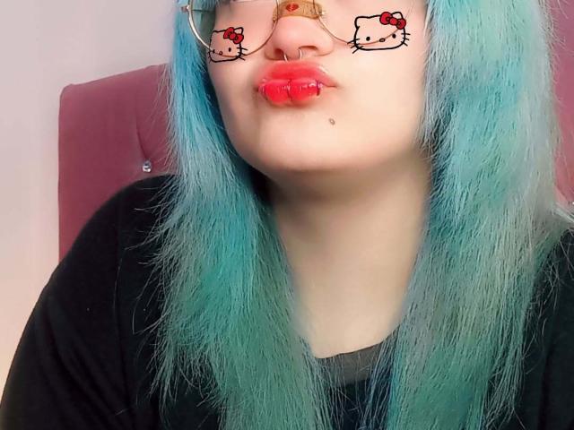 LuckyLeahh mit türkisfarbenem Haar, Brille mit Hello Kitty-Rahmen, rotem Lippenstift, Nasenring und Lippenring, leicht schmollend, schwarzes Shirt, helle Haut, süß, nerdig, sexy..