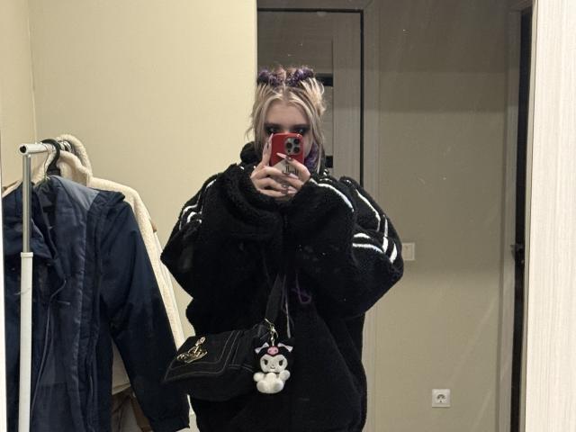 LennieCaronna with purple hair buns, black fuzzy jacket, black purse with stuffed toy, holding red phone, mirror selfie, beige room, blue jacket on hanger.
