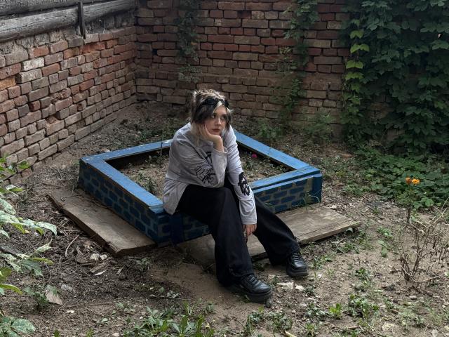 LennieCaronna with messy black-and-brown hair, sitting on a blue-bricked box, wearing a grey hoodie, black pants, and black boots, against a brick wall with green vines.