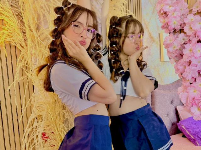 IsabelaP with curly brown hair, glasses, and a sexy Japanese schoolgirl outfit, pouting with hands on cheeks, mirror reflection showing same pose, pink cherry blossoms on the right.