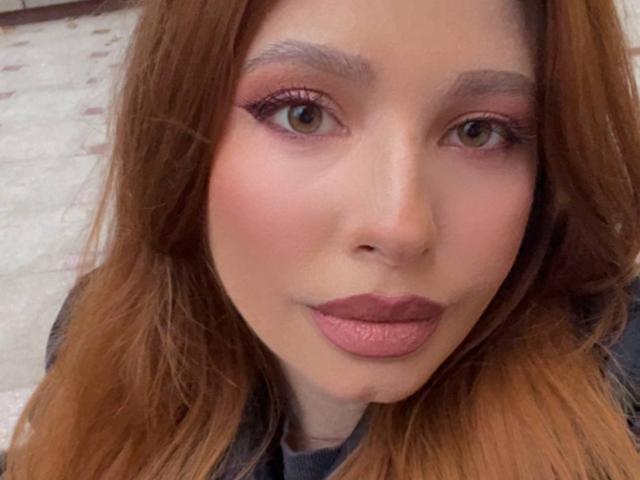 MiraVolt has brown eyes, pink eyeliner, and pink lipstick with her long brown hair down. She's looking at the camera with a slight smile, showing her full lips.
