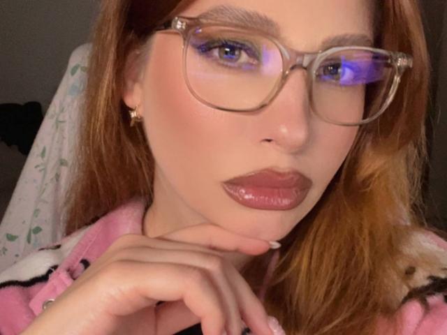 MiraVolt with glasses, pink lipstick, and shiny lips, holding her chin, wearing a pink top, has red hair and light skin. Her hand is near her face.
